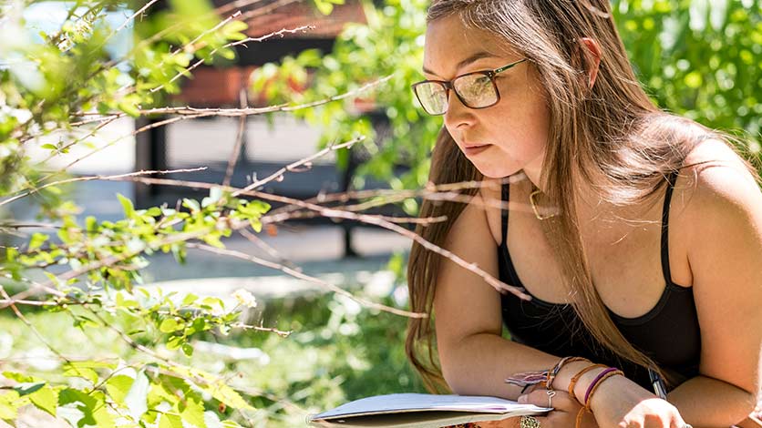 A young woman wearing a black tank top and glasses. She looks deep in thought and is standing somewhere outside. Tree branches can be seen in the foreground and background.