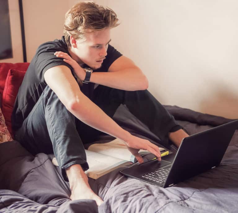 A young man sitting in an unmade bed. He's looking at his notebook computer.
