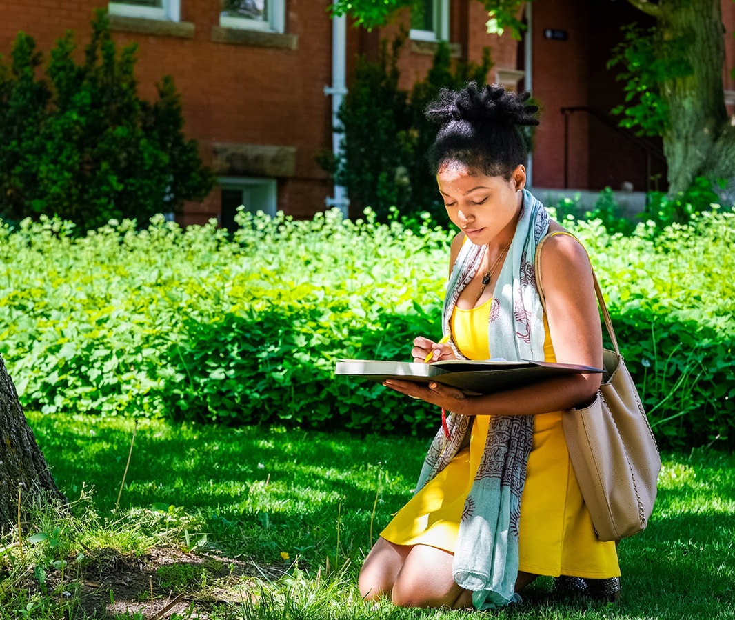 A young woman in a kneeling position holding a manuscript. She is outdoors in what appears to be a garden. In the background, the brick wall of a building can be seen.