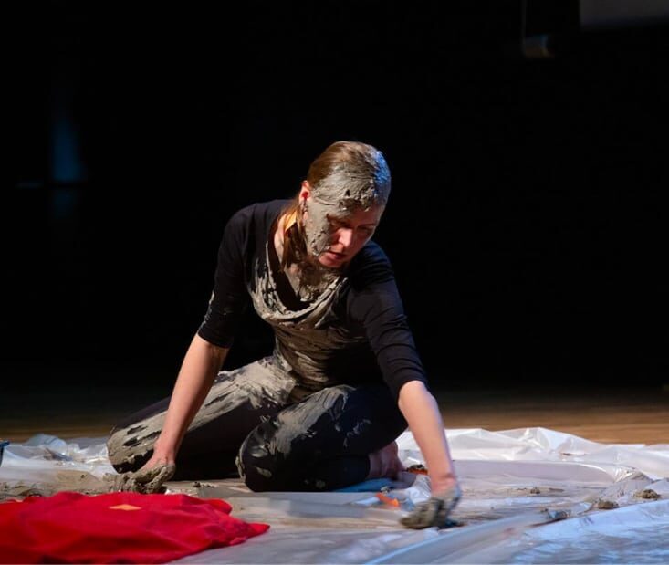 A woman is sitting on her knees and leaning forward and to the left with her torso. She's wearing black clothes which are partially stained with clay. There is clay all around her and on her hands and face. She seems to be experimenting with the material.