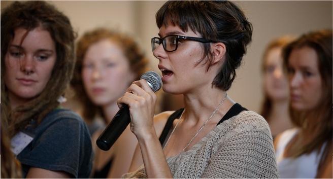 A woman holding a microphone and speaking. The picure shows her from the shoulders up. Other women are seen in the background, some looking at her and seemingly listening to what she's saying.