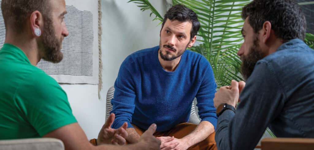 Three men sitting in a small circle, two men are intently listening as the third man speaks.