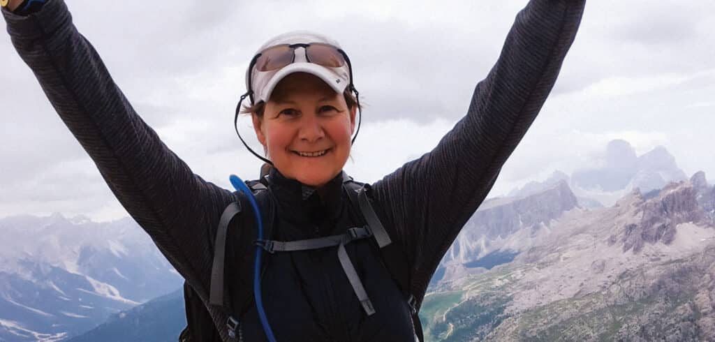 A woman standing in front of mountain peaks and clouds with both arms raised to the sky in victory.