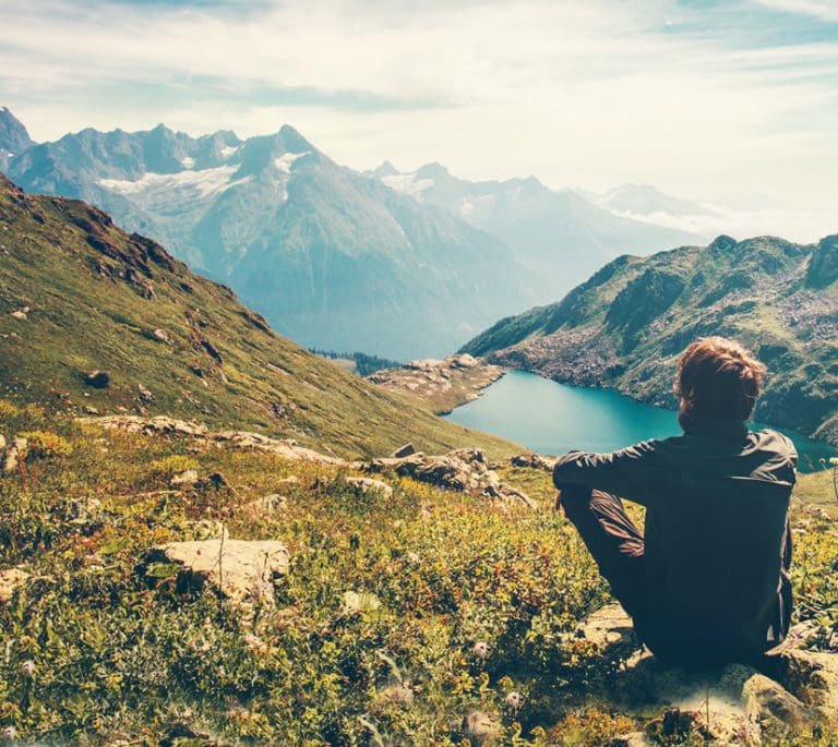A person is sitting on the ground looking out over the landscape, which is a lake surrounded by mountains.