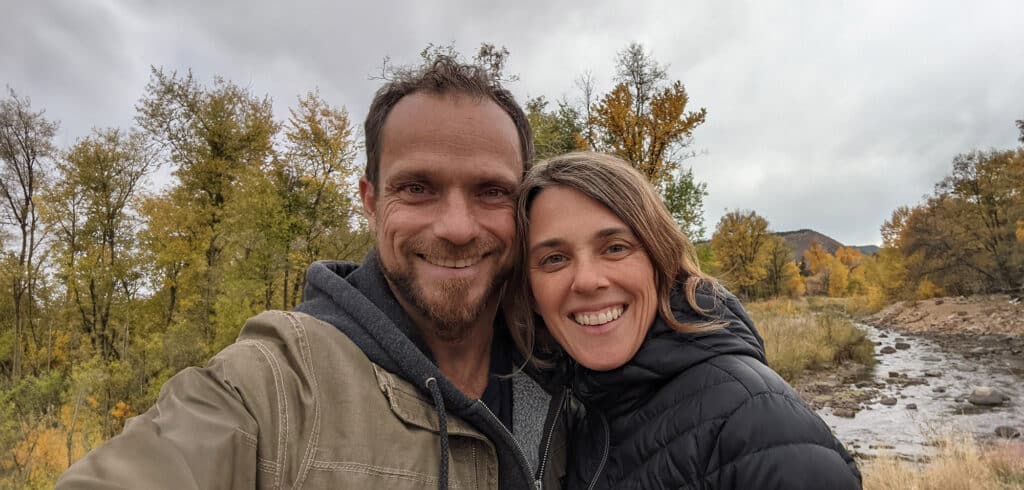 A man and woman snuggled together for a selfie with a creek, trees, and mountains in the background.