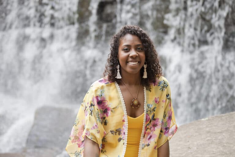 Kemba Douglas, beautiful black woman wearing a yellow shirt and yellow floral shawl, in front of a waterfall.