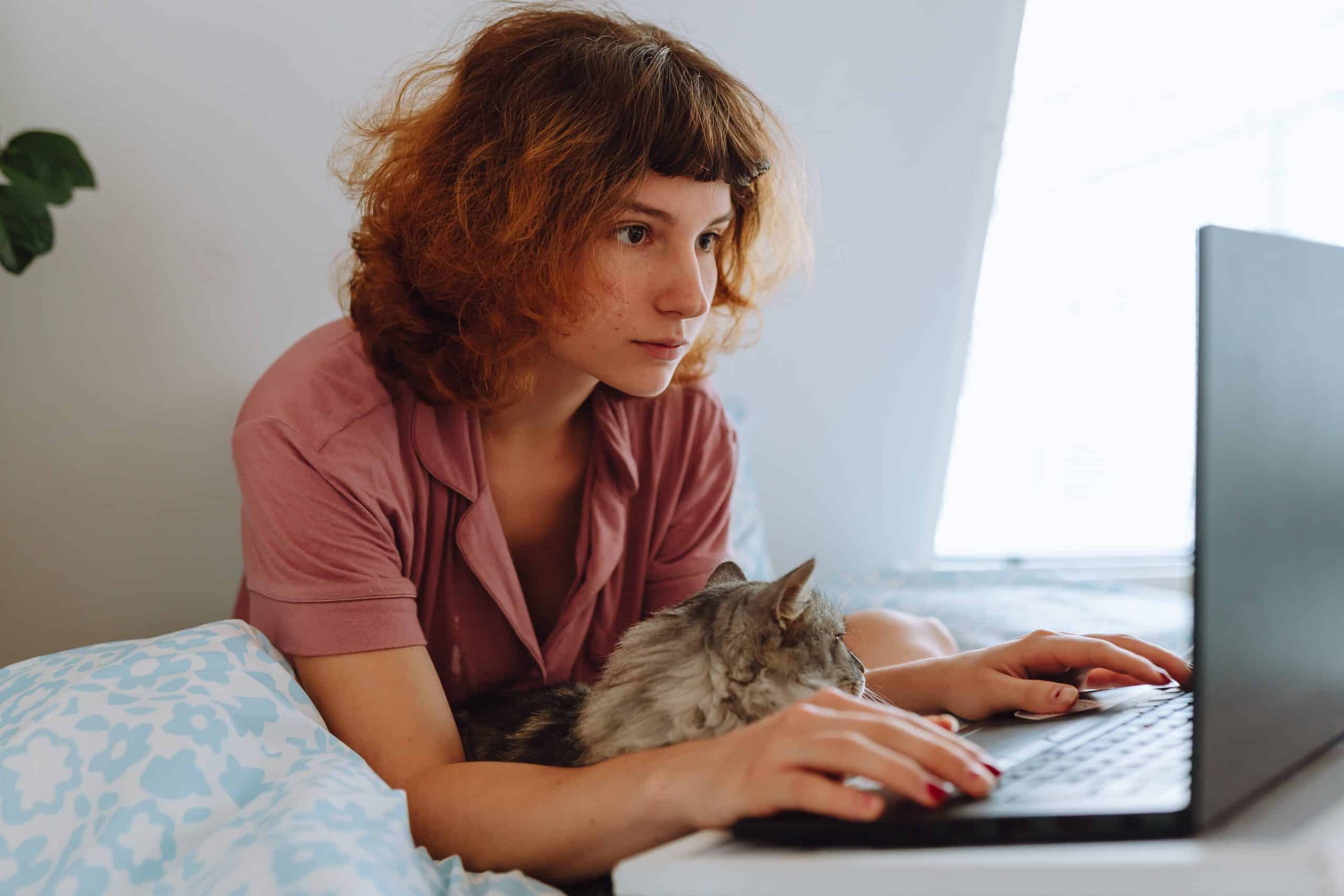 A woman sitting on a bed using a laptop with her cat on her lap