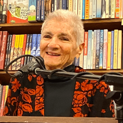 Margaret Randall at a microphone with books behind her