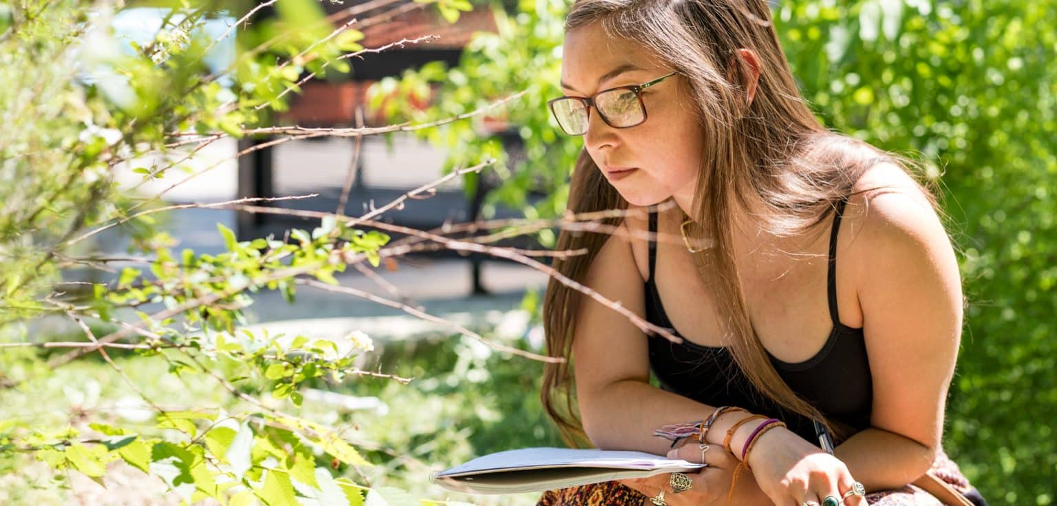 A woman in glasses holding a notebook looks closely at a green plant outside
