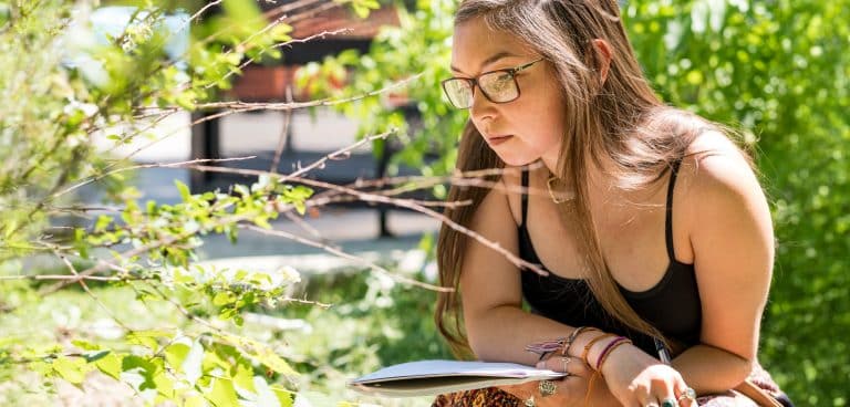 A woman in glasses holding a notebook looks closely at a green plant outside