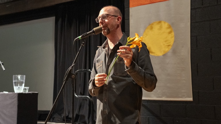 Jeffrey Pethybridge standing in front of a microphone holding a lily on stage in the Performing Arts Center.