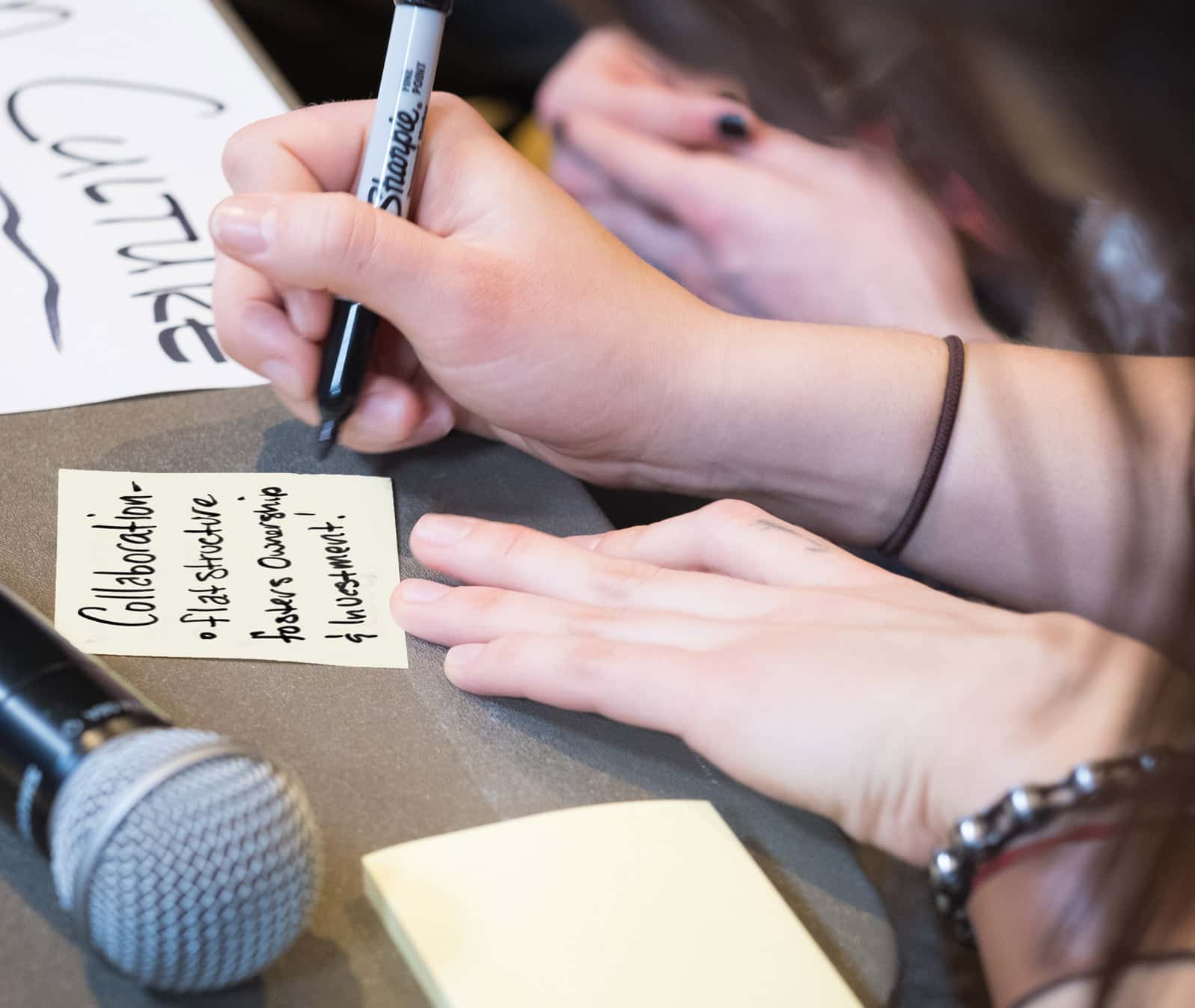 A close-up of a hand writing in a Sharpie marker on a sticky note.