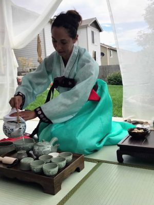 A woman in traditional Korean garb kneeling on a mat pouring tea in a tea ceremony