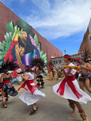 A group of people performing a traditional Aztec dance in front of a mural